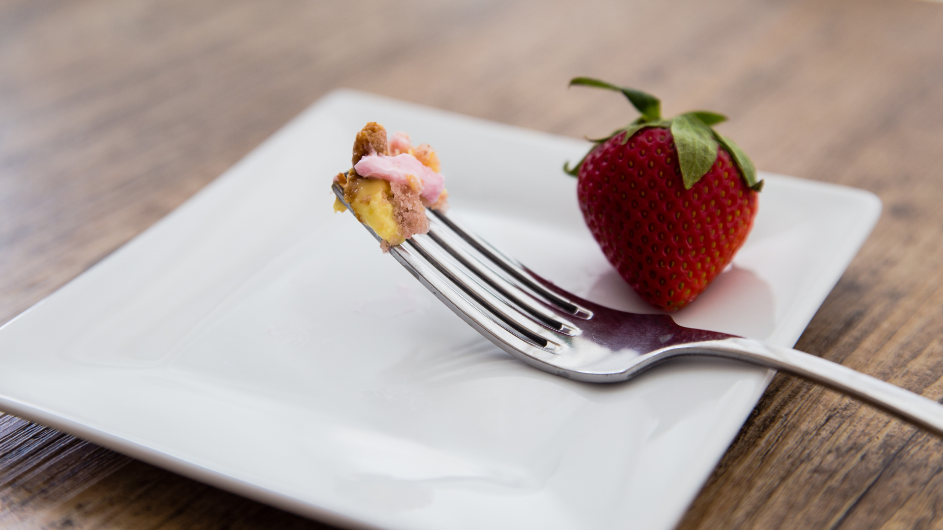 A small piece of cake on a fork resting on a small square white plate with a single strawberry.