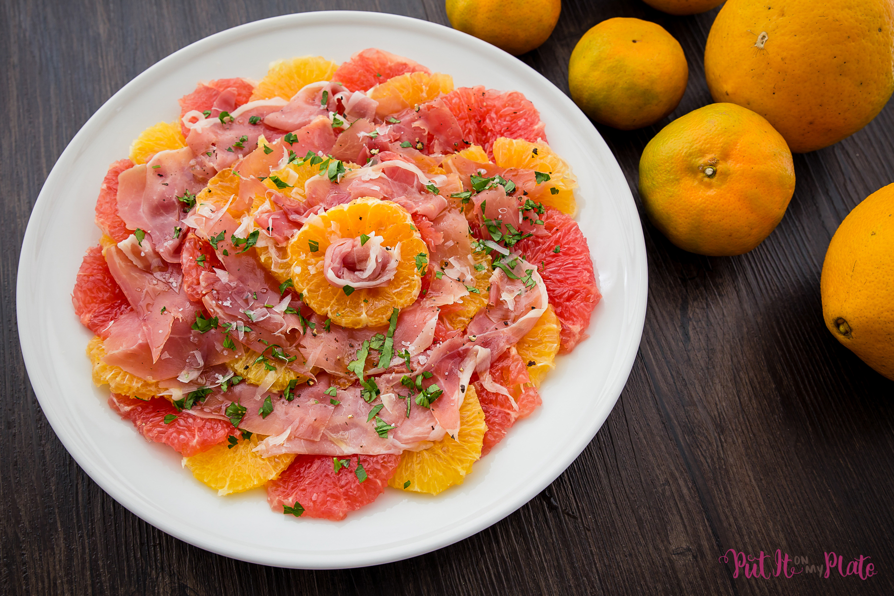 Local Citrus Salad - slices of oranges, tangerines and grapefruit laid in concentric overlapping circles on a white platter, with proscuitto and a sprinkle of chopped green herbs on top.