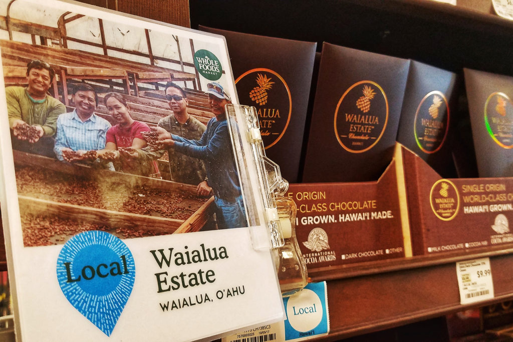 Photograph of Waialua Estate employees holding cacao beans used on a Whole Foods shelf sign in front of retail chocolate bars for sale.