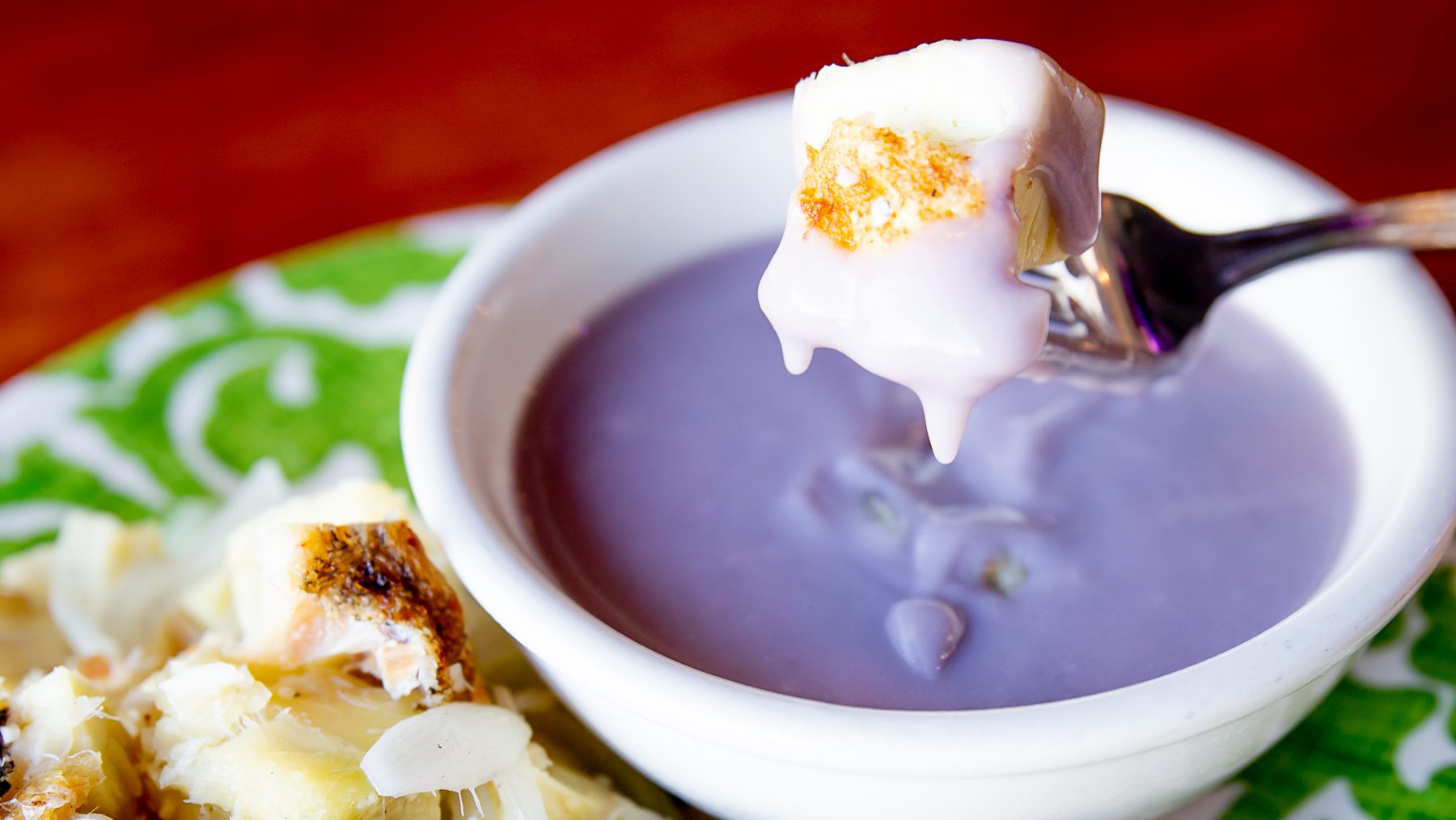 A small piece of fried breadfruit dipped in poi being held up on a fork over a white bowl full of poi, with fried breadfruit and onions on the side.