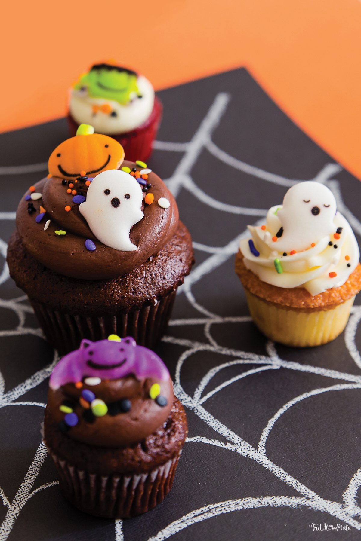 Cake Works Bakery - Halloween Cupcakes - assorted miniature cupcakes with monster sugar decorations sitting on a cobweb design paper.