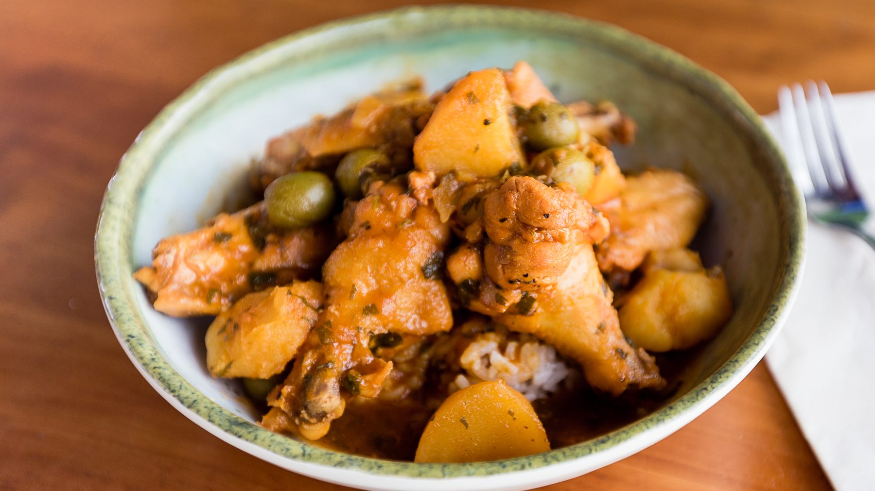 A shallow pale green ceramic bowl filled with cooked chicken drumettes, potato chunks and green olives on a bed of rice.