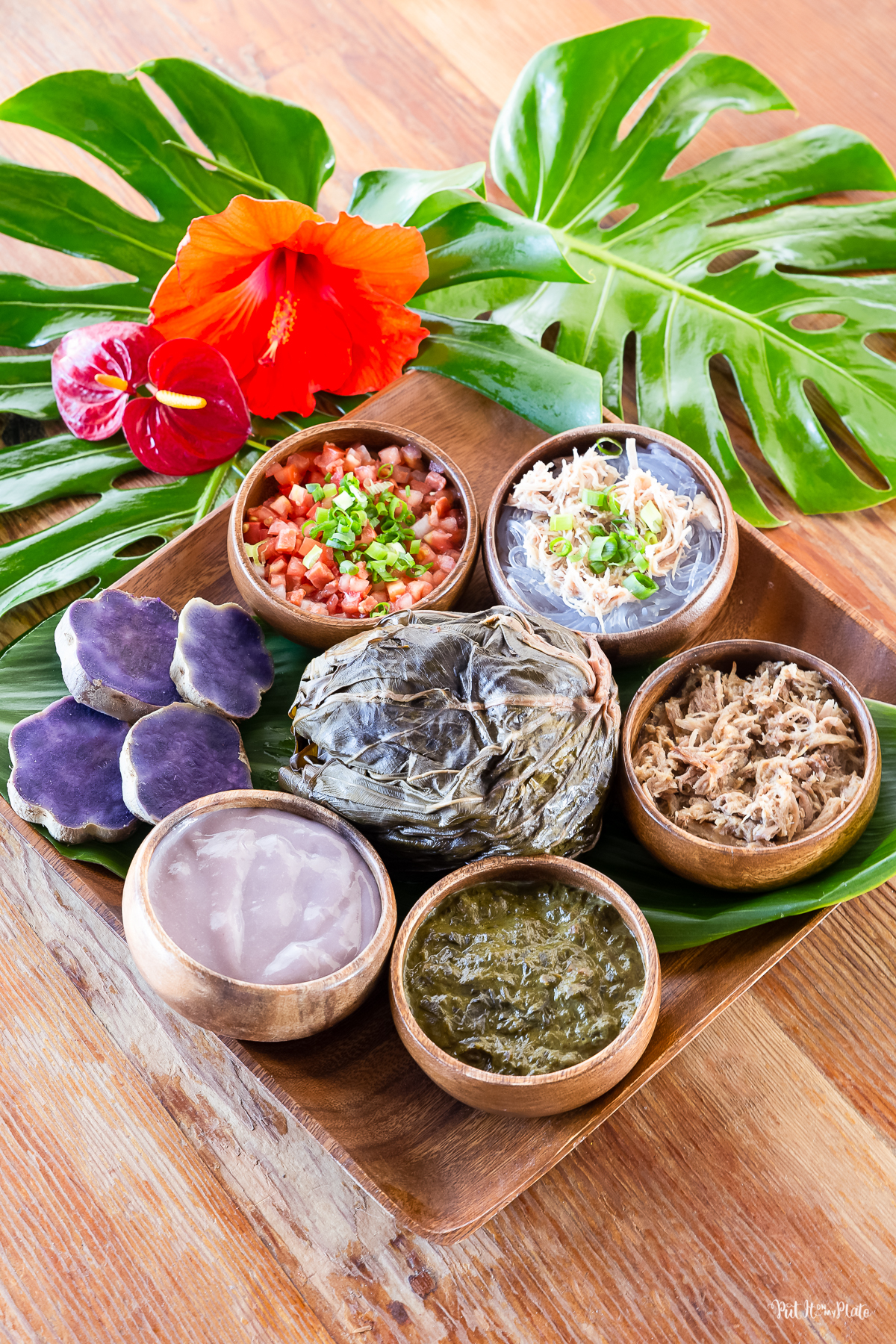 Hawaiian Food Sampler - wooden bowls of poi, squid luau, kalua pig, chicken long rice and lomi salmon arranged on a wooden plate around a pork lau lau, with four pieces of purple sweet potato.